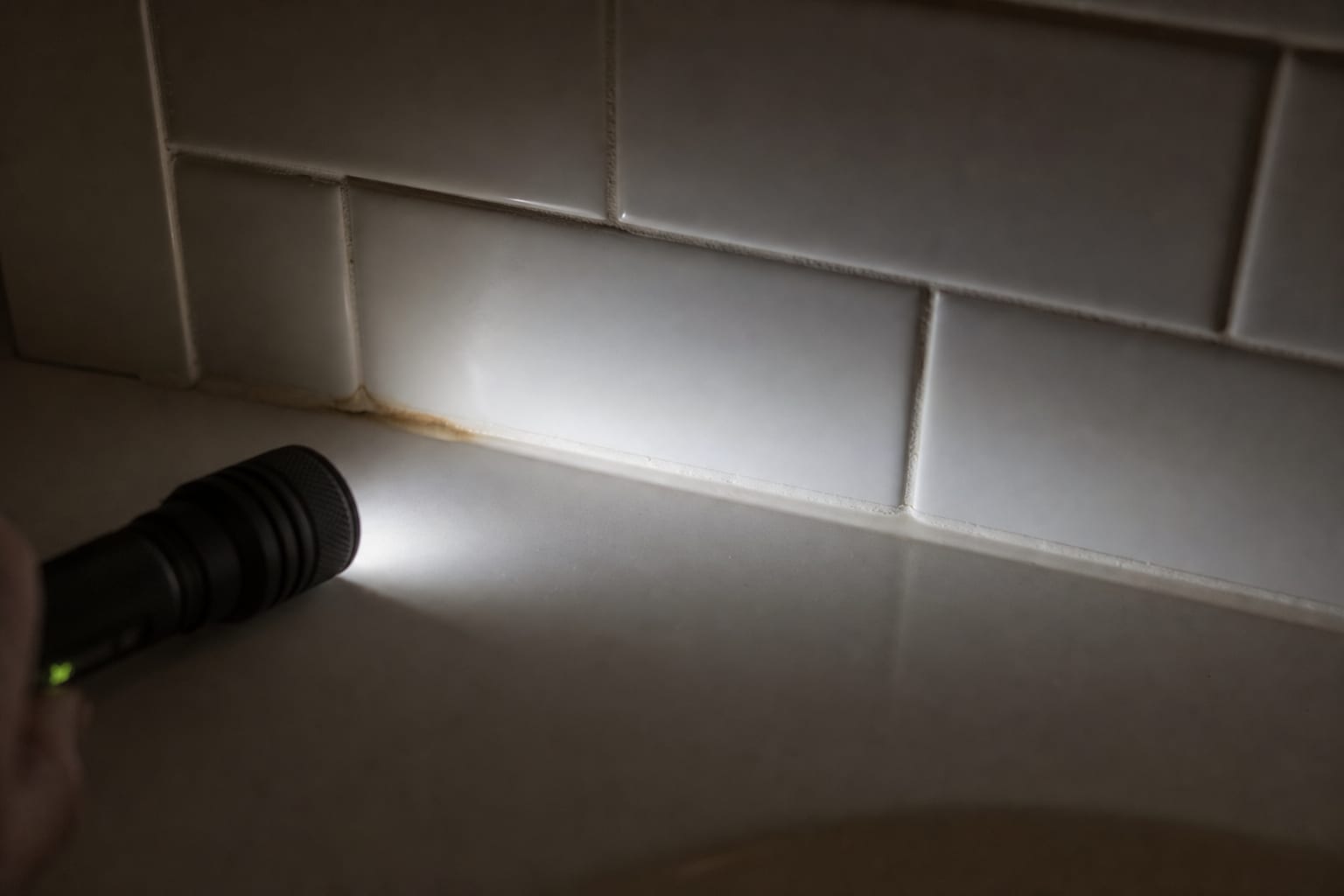 Close-up of the joint where white subway tile meets a bathtub deck in a residential bathroom, sealant bead visible along the seam with slight discoloration a...